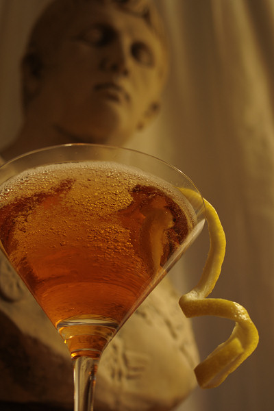 photo of a cocktail in a martini glass with a lemon twist in front of a bust of Augustus Caesar